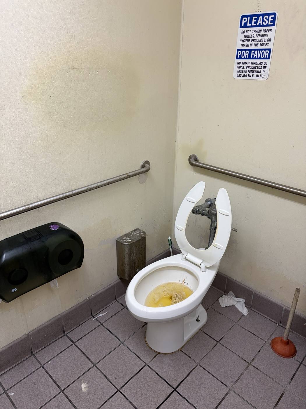 An accessible restroom stall: unflushed toilet with raised seat, rusted sanitary bin, empty dual-roll dispenser, plunger in the corner, bilingual sign on the wall
